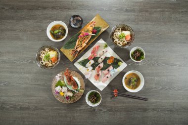Çeşit çeşit suşi, ızgara karides, udon eriştesi ve ahşap bir masaya yerleştirilmiş garnitür içeren tam bir Japon yemeği..