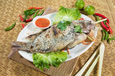 Marul yapraklarıyla servis edilen ızgara bir balık, kırmızı biberli garnitür ve desenli bir paspasın üzerinde sos..