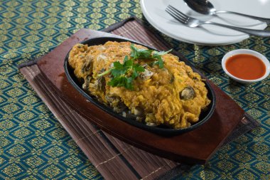 Siyah bir sıcak tabakta cızırdayan bir istiridye omleti kişniş ile süslenmiş ve yanında biber sosu ile desenli bir paspas..