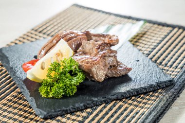 Limon dilimi ve maydanozla servis edilen ızgara sığır dili dilimli siyah bir tabak..