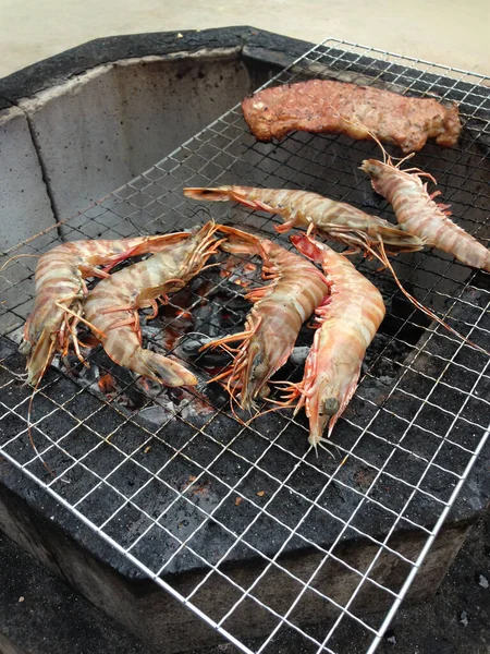 Birkaç büyük taze karides ve bir parça bifteğin açık hava barbekü ocağında ızgarada pişirildiği yakın bir yatay fotoğraf. Görüntü, deniz ürünlerinin canlı kırmızı ve turuncu renklerini ve metal tel askıdaki eti yakalıyor.,