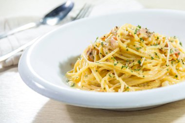Arka planda çatal ve kaşıkla servis edilen, üzerinde otlar ve küçük pastırma parçaları olan kremalı spagetti ile dolu beyaz bir tabak..