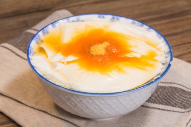 Üzerinde altın şurup ve şeker tozu bulunan pürüzsüz soya peyniri ile dolu beyaz bir seramik kase çizgili bir kumaşa konur..