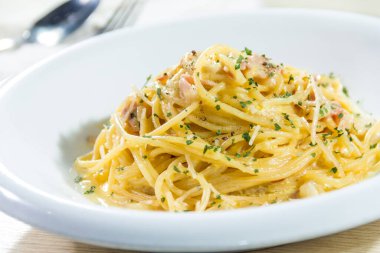 Arka planda çatal ve kaşıkla servis edilen, üzerinde otlar ve küçük pastırma parçaları olan kremalı spagetti ile dolu beyaz bir tabak..