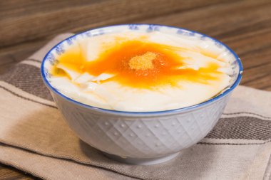 Üzerinde altın şurup ve şeker tozu bulunan pürüzsüz soya peyniri ile dolu beyaz bir seramik kase çizgili bir kumaşa konur..