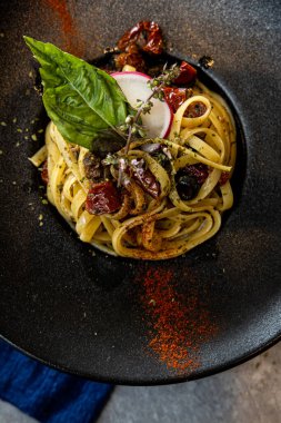 Fesleğen yaprakları, güneşte kurutulmuş domatesler ve kırsal bir Akdeniz sunumu için otlarla süslenmiş siyah seramik bir tabak..