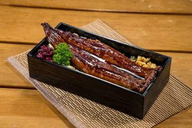 Soya soslu ızgara yılan balığı filetosu ile kaplı, maydanozla süslenmiş beyaz pirinçle dolu dikdörtgen bir bento kutusu..