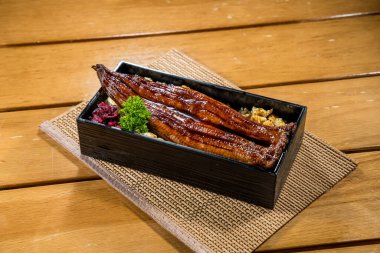 Beyaz pirinçle dolu dikdörtgen siyah bento kutusu üzerine ızgara yılan balığı filetosu, tatlı soya sosuyla kaplanmış, maydanozla süslenmiş..