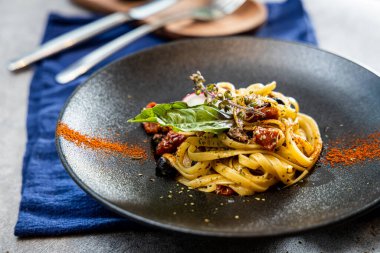 Mavi bir kumaş üzerinde servis edilen taze fesleğen yaprakları, kiraz domatesleri ve otlarla süslenmiş İtalyan makarnasıyla dolu siyah bir seramik tabak..