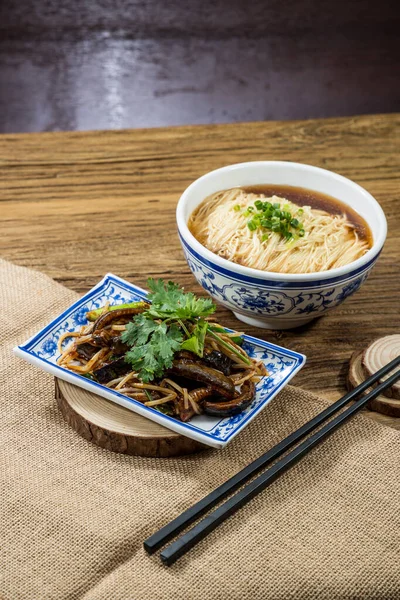 Soya bazlı çorbada ince erişteyle dolu mavi desenli bir kase kişniş ve yeşil soğanla süslenmiş bir tabak haşlanmış yılan balığı..