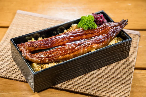 Soya soslu ızgara yılan balığı filetosu ile kaplı, maydanozla süslenmiş beyaz pirinçle dolu dikdörtgen bir bento kutusu..