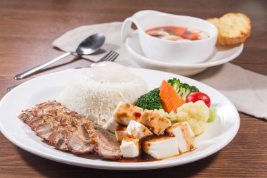 Buharda pilav, soslu tofu küpleri, ızgara et dilimleri ve yanında çorba ve ekmekle servis edilen çeşitli sebzelerden oluşan beyaz bir tabak..