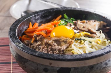 Üzerinde çeşitli sebzeler, sığır eti dilimleri, mantarlar ve ortasında çiğ yumurta sarısı olan buğulanmış pirinçle dolu siyah taş bir kase..