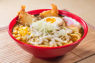 Tempura karidesi, haşlanmış yumurta, mısır ve dilimlenmiş sebzelerle kaplı kırmızı bir udon eriştesi..
