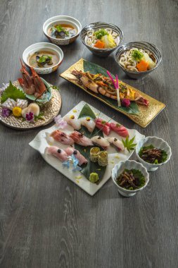 Zarif Japon seti içinde udon kaseleri ve temiz çorba, çeşitli bir tabak nigiri ve büyük bir ızgara karides var. Süslemeler ve mevsimlik dokunuşlar renk ve denge sağlar. Bu da çok yönlü, düşünceli bir yemek olduğunu gösterir..