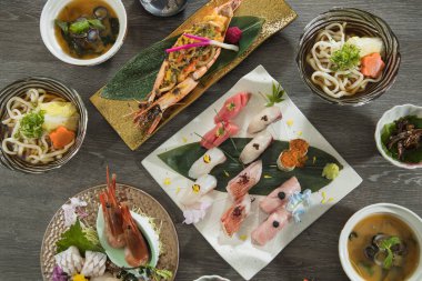 İnce dilimlenmiş çiğ balık ön planda yapraklarla süslenmiş bir şekilde duruyor. Arkasında yeşil, mantar, tofu ve kök sebzeleri küçük daldırma kapları olan bambu bir hasırın üzerinde saklar..