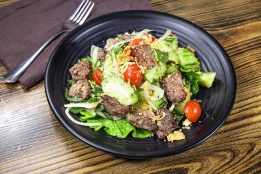 Karma yeşillikler, kiraz domatesleri, soğan dilimleri, şifalı otlar ve ızgara et küpleriyle dolu siyah tabak. Hafif soslar ve kıtır kıtır dokular sağlıklı öğle yemeği menüleri ve kafe promosyonları için protein açısından zengin bir salata öneriyor..