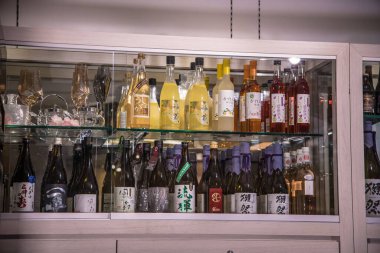 Arka plandaki cam dolapta iki rafta geniş çaplı sake şişeleri bulunuyor. Japon etiketli temiz ve kehribar şişeleri kristal bardakların ve bar eşyalarının yanında duruyor. Bu da bir dükkan veya bar ortamında rafine edilmiş bir tadımlık yeri işaret ediyor..