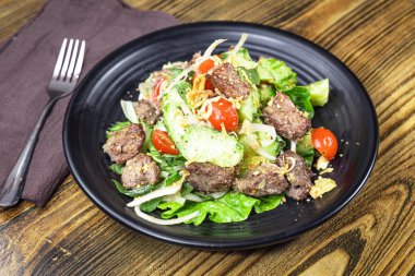 Siyah tabakta kurutulmuş sığır eti parçaları, kıtır marul, vişneli domates ve kızarmış soğanlı canlı bir salata var. Kırsal ahşap masa ve keten peçete sıradan bir bistro tarzı yemek olduğunu gösteriyor..