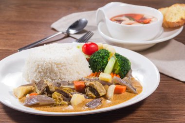 Körili sığır eti, çeşitli sebzeler ve ahşap bir masada bir kase çorba ile servis edilen bir tabak beyaz pirinç.