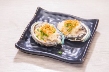 İki deniz kulağı kabuğu, üzerinde sarımsak ve çıtır ekmek kırıntıları var. Siyah bir tabakta servis ediliyor..