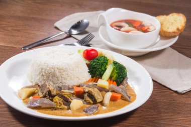 Tayland usulü körili biftek, sebze ve bir kase çorbanın yanında, buğulanmış pirinçli beyaz bir tabak..