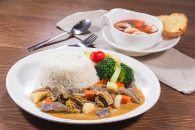 Tayland usulü körili biftek, sebze ve bir kase çorbanın yanında, buğulanmış pirinçli beyaz bir tabak..