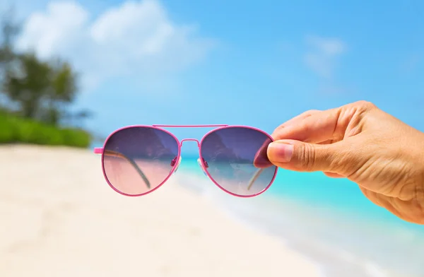 Una mano che tiene gli occhiali da sole su uno sfondo bellissimo spiaggia tropicale — Foto Stock