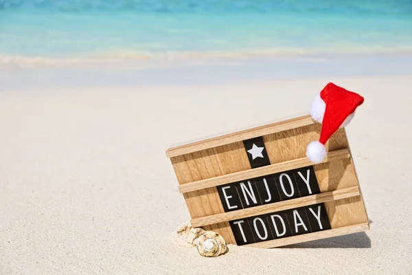 Wooden board with inspiration text ENJOY TODAY on white sandy beach, with small red Santa hat, copy space