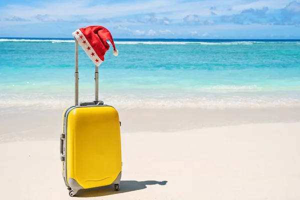 Red Santa Claus Christmas hat on handle of yellow travel luggage with tropical beach and sea — Stock Photo, Image