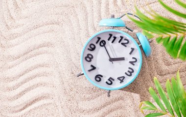 Kum arka planında mavi alarm saati, kopyalama alanı ve yeşil palmiye yapraklarıyla üst görünüm