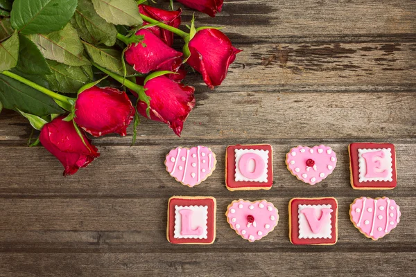 Red roses and pink cookies in shape of LOVE word, on wooden back — Stock Photo, Image