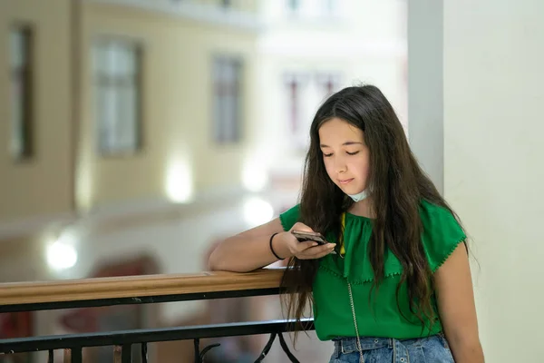 Kız balkonda duruyor ve telefona mesaj yolluyor. Yeşil bluzlu bir kız, alışveriş merkezinin balkonunda yüzünde koruyucu bir maskeyle duruyor. Yeni bir gerçeklikte koruma kavramını kabul et