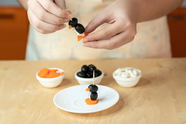 Kadınların elleri sağlıklı bir yemek hazırlıyor. Zeytin, kaymak peyniri ve penguen şeklinde havuçtan yapılmış şiş aperatifler. Adım 3