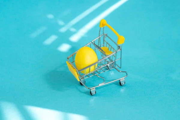 Mavi arka planda süpermarket tramvayındaki sarı paskalya yumurtası. Tatil alışverişi konsepti