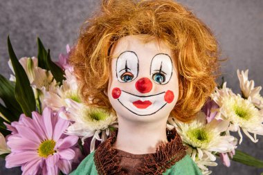 Turuncu saçlı, kırmızı burunlu, bahar çiçekleriyle çevrili antik palyaço bebeği.