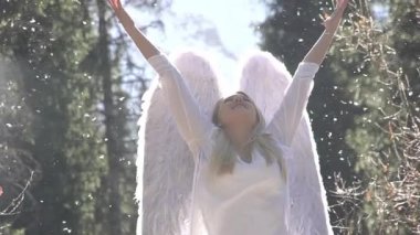 Güneşin ışınlarındaki neşeli melek gibi. Beyaz elbiseli güzel bir sarışın ellerini havaya kaldırıyor. Arkasında beyaz kanatları var. Havada beyaz tüyler uçuşuyor. Slow Motion 240 fps hızında