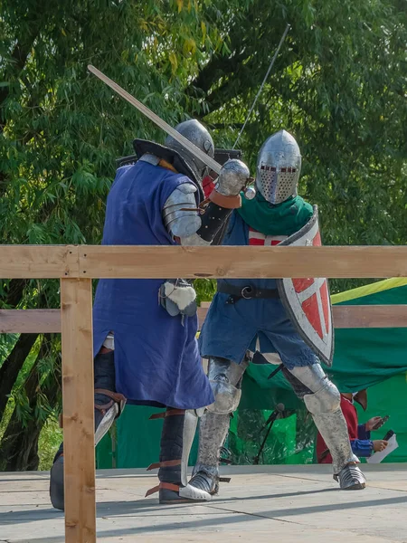 Two foot warriors in heavy medieval armor fight in the arena. Armed with swords. They are protected by iron helmets and shields. Historical reconstruction of medieval European knightly tournaments