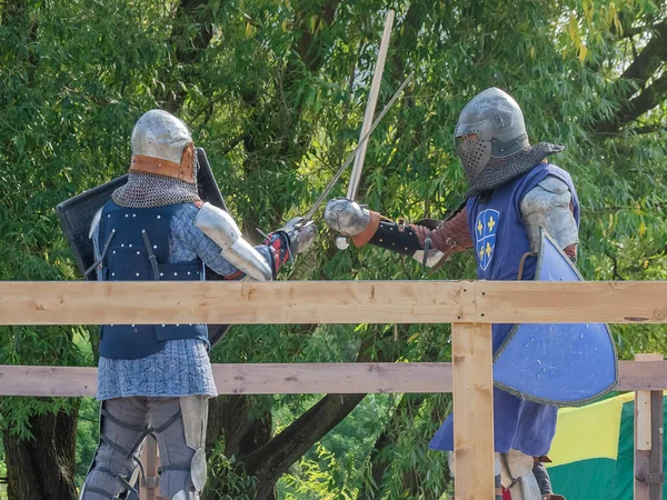 Two foot warriors in heavy medieval armor fight in the arena. Armed with swords. They are protected by iron helmets and shields. Historical reconstruction of medieval European knightly tournaments