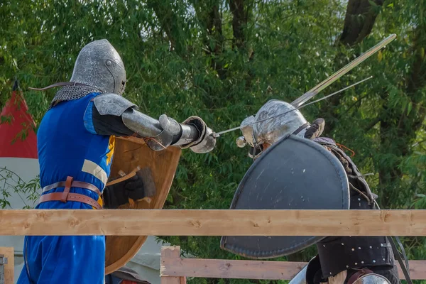 重中世の鎧を着た2人の足の戦士がアリーナで戦う 剣で武装してる 鉄のヘルメットとシールドで保護されています 中世ヨーロッパ騎士大会の歴史的復興 — ストック写真