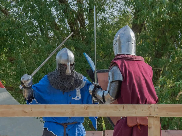 Two foot warriors in heavy medieval armor fight in the arena. Armed with swords. They are protected by iron helmets and shields. Historical reconstruction of medieval European knightly tournaments