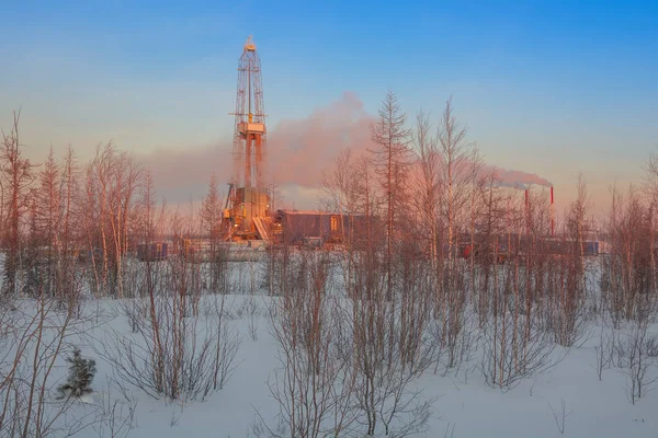 A rig for drilling oil and gas wells in the northern landscape of the forest-tundra. The rising winter sun colors the landscape orange. Puffs of steam and smoke partially cover the oil derrick