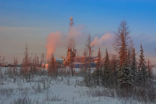 A rig for drilling oil and gas wells in the northern landscape of the forest-tundra. The rising winter sun colors the landscape orange. Puffs of steam and smoke partially cover the oil derrick