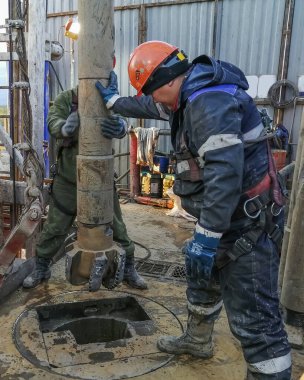 Nadym Rusya - 12 Haziran 2021: İki Asyalı sondaj işçisi kayaları delmek için petrol ve gaz kuyusuna PDC ucu yerleştirdi. Özel kıyafetler giyiyorlar, koruyucu miğfer takıyorlar. Teçhizatın içinde.