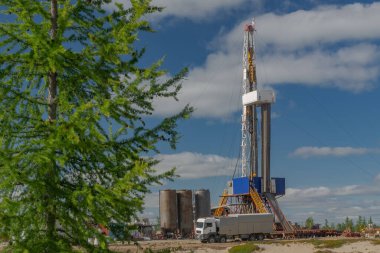 Petrol ve gaz sahasında sondaj kulesiyle manzara. Altyapı, iletişim ve sondaj ekipmanları görülebilir durumda. Platformun etrafında yeşil ağaçları ve mavi gökyüzü olan kuzey taygası var.