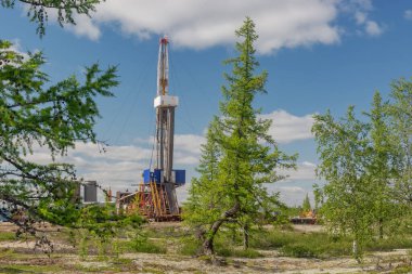 Petrol ve gaz sahasında sondaj kulesiyle manzara. Altyapı, iletişim ve sondaj ekipmanları görülebilir durumda. Platformun etrafında yeşil ağaçları ve mavi gökyüzü olan kuzey taygası var.