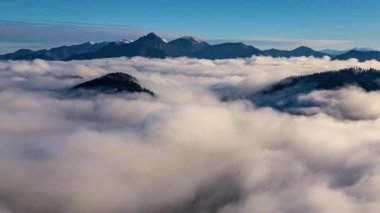 Gün doğumunda dağlarda süzülen puslu bulutlar. Sıcak güneş ışığı doğal manzarayı aydınlatırken sisli bulutlar mavi bir gökyüzünün altında huzur içinde süzülüyor ve sakin ve manzaralı bir manzara yaratıyor. Havadan