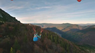 Güneşli dağlardaki renkli sonbahar ormanlarının üzerinde özgürce süzülen bir paraşütçünün sakin akşam uçuşu. Açık hava macerasının adrenalinini yakalamak, huzurlu bir eğlence ve özgür olma hissi.