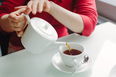 Woman pouring black tea from a teapot into a white cup, close-up.