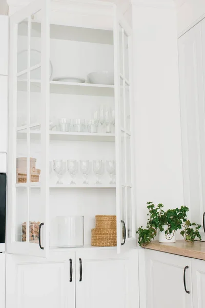 Opened white glass cabinet with clean dishes and decor. Scandinavian style kitchen interior. Organization of storage in kitchen. Vertical photo.